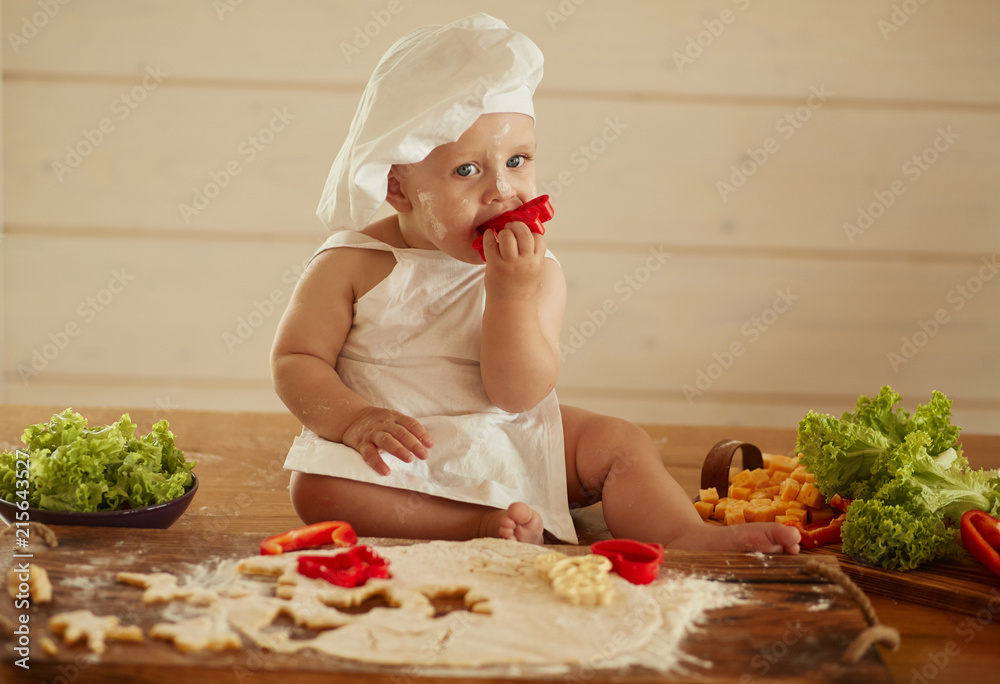 The small child sits on the table near dough and vegetables The small child sits on the table near dough and vegetables