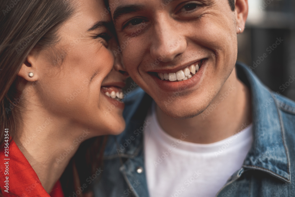 Close up of male and female smiling faces. Loving couple is standing with delight and embracing. Affectionate girl is kissing boyfriend and laughing Close up of male and female smiling faces. Loving couple is standing with delight and embracing. Affectionate girl is kissing boyfriend and laughing