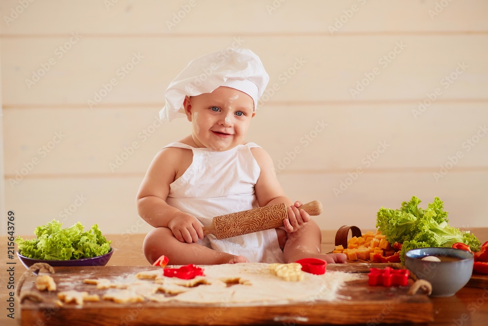 The small child sits on the table near dough and vegetables The small child sits on the table near dough and vegetables