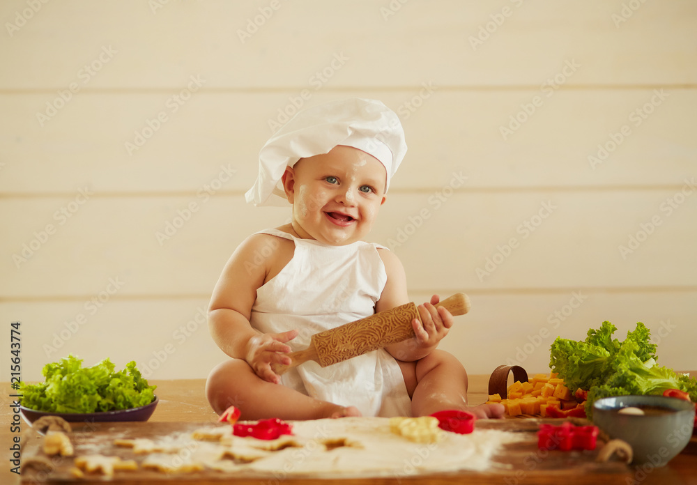 The small child sits on the table near dough and vegetables The small child sits on the table near dough and vegetables