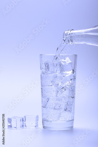 Pouring fresh water into glass with ice from a bottle. Ice cubes next to glass.
