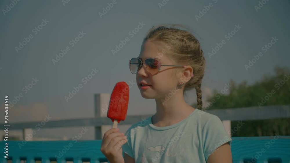 Portrait of lovely happy preteen girl in sunglasses eating delicious red fruit ice cream on stick in summertime while enjoying free time in park. Beautiful child eating popsicle outdoors. Slo mo. Stock ビデオ | Adobe Stock