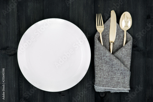 a set of bronze cutlery  spoon  fork and knife wrap with gray napkin placing beside white plate on black wooden table