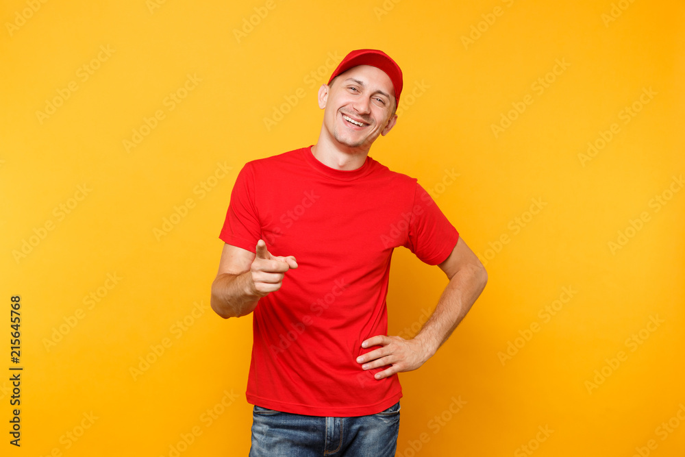 Delivery man in red uniform isolated on yellow orange background. Professional male employee in cap, t-shirt working as courier or dealer pointing index finger camera. Service concept. What about you