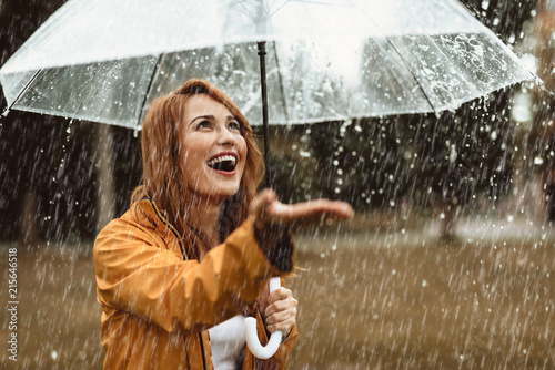 Waist up portrait of smiling girl standing outdoors with umbrella in hands. She is stretching hand and opening mouth in true pleasure