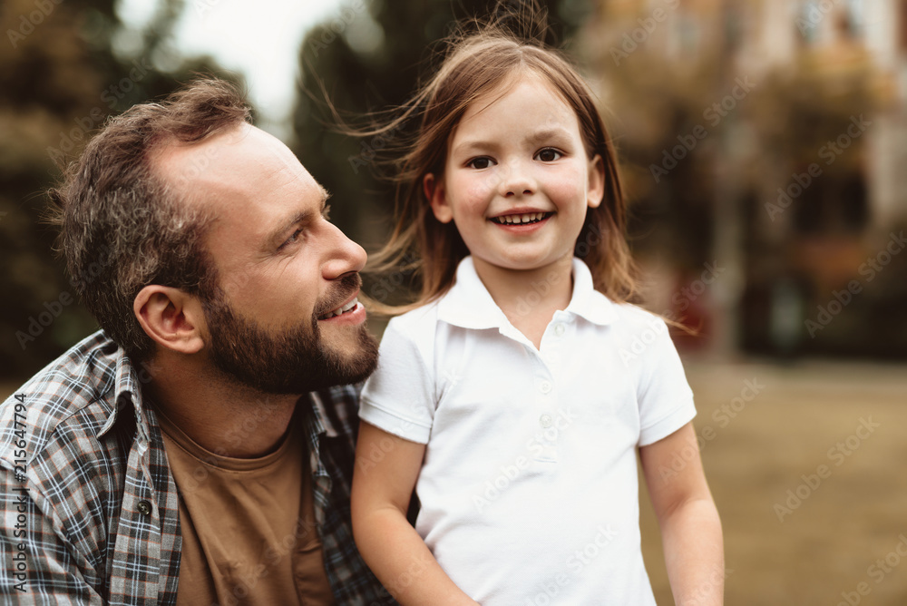 Waist up portrait of happy daughter and dad enjoying time outdoors. Small kid is standing by delighted man squatting near child and looking at her with joy