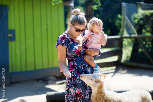 Adorable cute toddler girl and young mother feeding little goats and sheeps on a kids farm. Beautiful baby child petting animals in the zoo. woman and daughter together