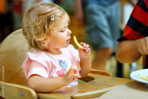 Adorable toddler girl eating healthy vegetables and unhealthy french fries potatoes. Cute happy baby child taking food from dish at daycare or nursery canteen.