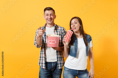 Young smiling couple woman and man watching movie film on date holding bucket of popcorn plastic cup of soda or cola pointing index finger isolated on yellow background. Emotions in cinema concept.