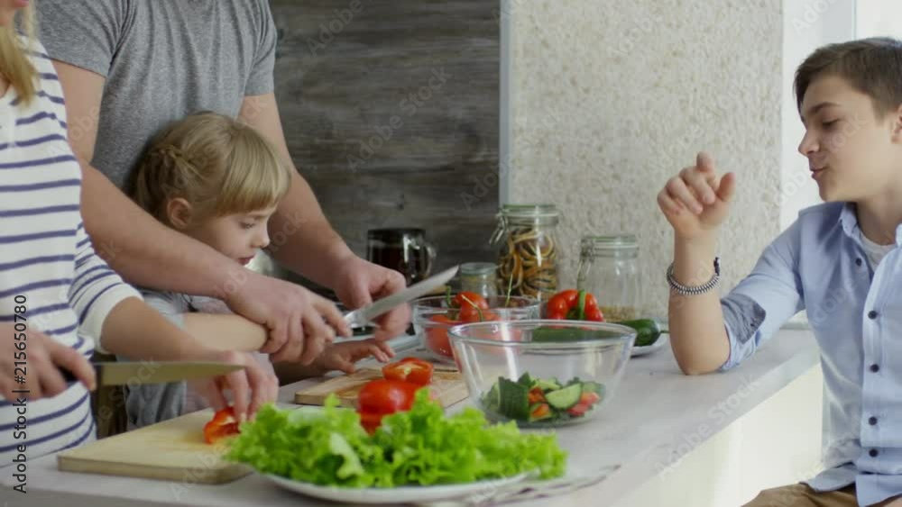 Tracking shot of mother, father and little daughter cutting vegetables for salad together while son sitting at kitchen counter and telling something to family Tracking shot of mother, father and little daughter cutting vegetables for salad together while son sitting at kitchen counter and telling something to family