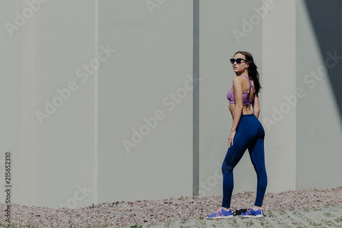 Full length shot of sporty slim woman with appealing look, stands sideways, wears shades, leggings and top, going to have outdoor workout, likes sport and gymnastics. People and aerobis concept photo