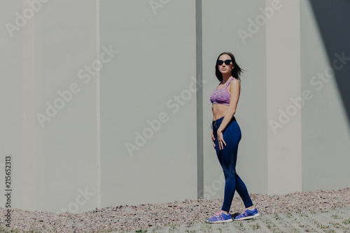 Horizontal shot of active fit woman in sportsclothes looks confidently into distance, wears shades, has rest after good outdoor exercise, cares of her health and figure. People, healthy lifestyle
