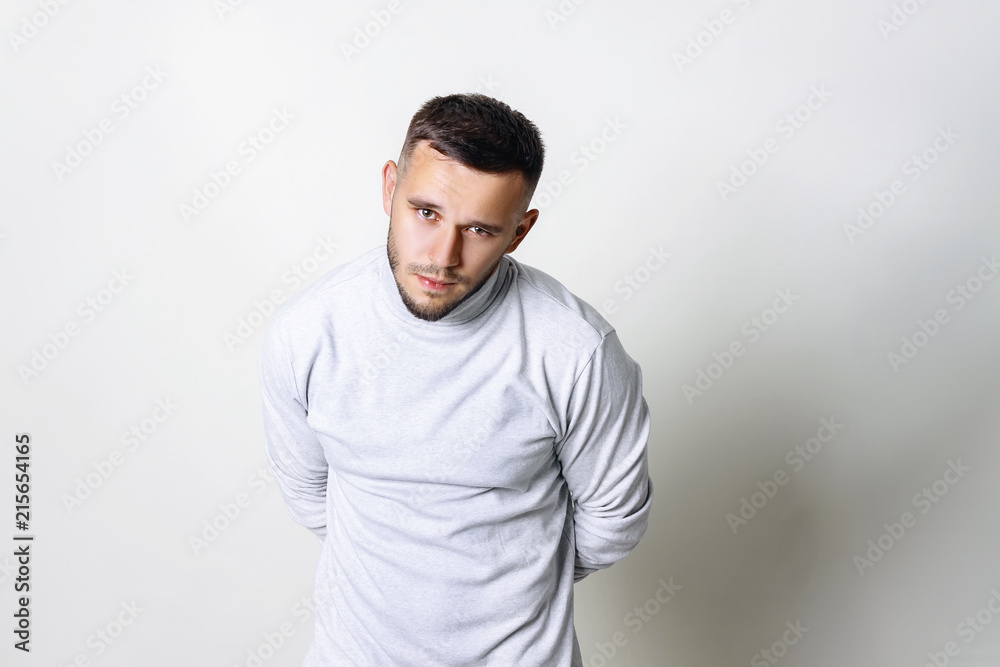 Fototapeta premium Close-up portrait of a sad man. Resentment of a man in white turtleneck his hands behind his back and lowered his head on grey background copy space