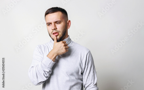 Handsome young man touching lips with his thumb and looks to side. Human emotions man in white turtleneck on grey background copy space © oleg_ermak