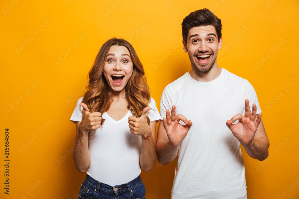 Photo of excited coup;e man and woman in basic clothing screaming and gesturing symbols with fingers at camera, isolated over yellow background Photo of excited coup;e man and woman in basic clothing screaming and gesturing symbols with fingers at camera, isolated over yellow background