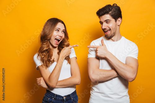 Image of happy young people man and woman in basic clothing laughing and pointing fingers at each other, isolated over yellow background