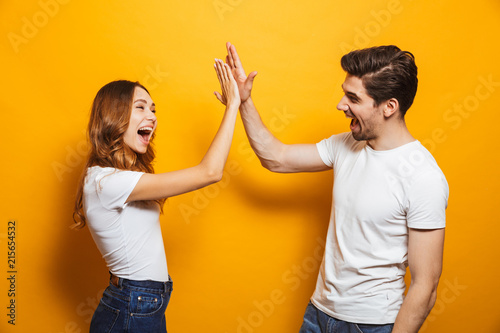 Image of friendly young people man and woman in basic clothing laughing and giving high five, isolated over yellow background photo