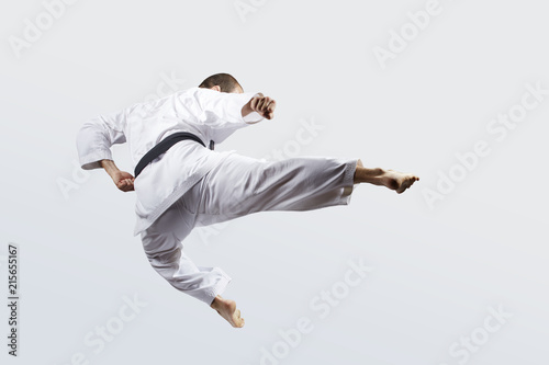 On a light background, an adult athlete with a black belt beats a kick in jump