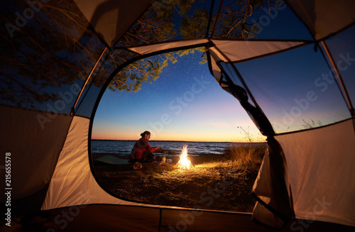 Camping on sea shore at night. View from tourist tent young woman backpacker preparing food on campfire on dark blue evening starry sky and clear water background. Tourism and adventure concept.