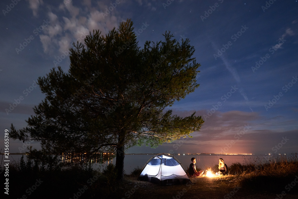 Small tourist tent under shadowy big tree and young couple, bearded man and pretty woman sitting at night on lake shore by bonfire under blue evening sky. Tourism, camping and loving relations concept