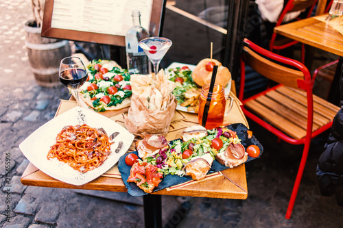 Restaurant table setting with food outdoor on the street. Italian pasta spaghetti, glass of red wine, mozarella cheese, bread, salad, tomatoes, burger and meat. Top view.