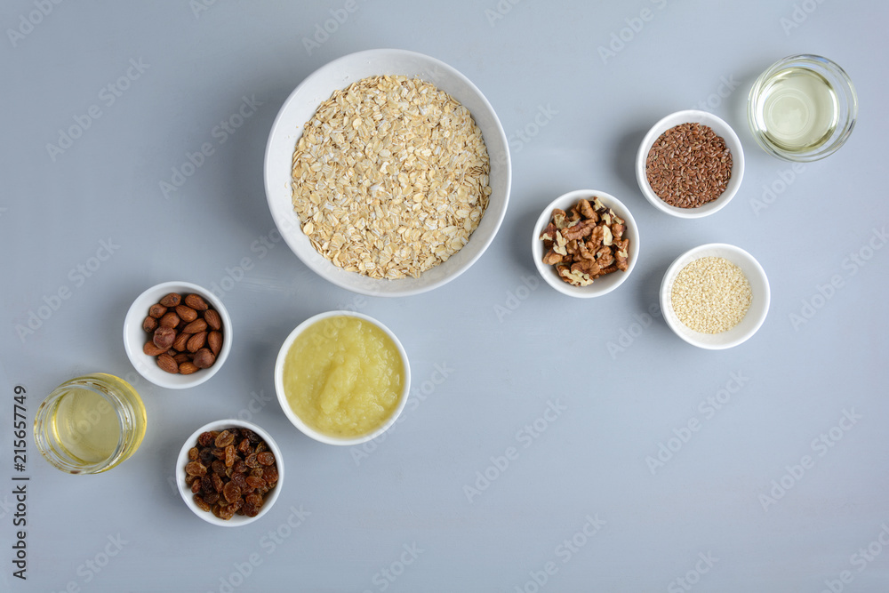Ingredients for cooking homemade granola: oatmeal, applesauce, honey, oil, almond, walnut, raisin, flax and sesame seeds on wooden gray background. Flat lay. Top view. Ingredients for cooking homemade granola: oatmeal, applesauce, honey, oil, almond, walnut, raisin, flax and sesame seeds on wooden gray background. Flat lay. Top view.