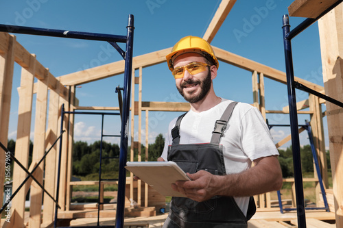 The man is a builder on the background of the roof of a frame house, in a yellow helmet and gray overalls. Uses a tablet photo