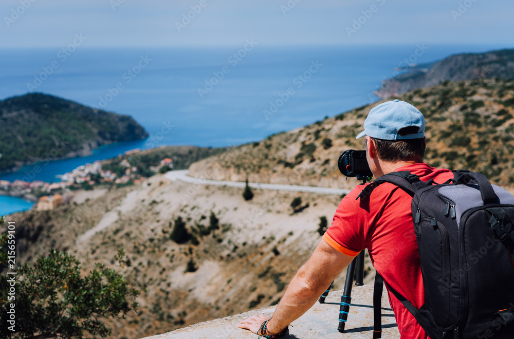 Male summer holiday vacation on Kefalonia Greece. Photographer with backpack enjoying capture of Mediterranean village Assos from top view point. Camera on tripod