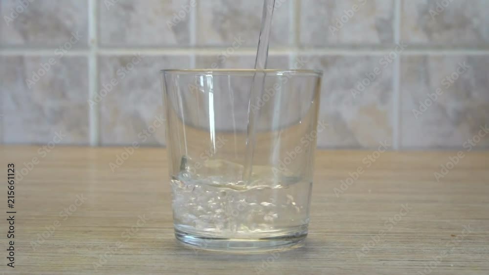 Transparent glass Cup. Water pours from above. Kitchen interior. Light background. Wooden table. Square tiles on the wall. Quenching thirst.