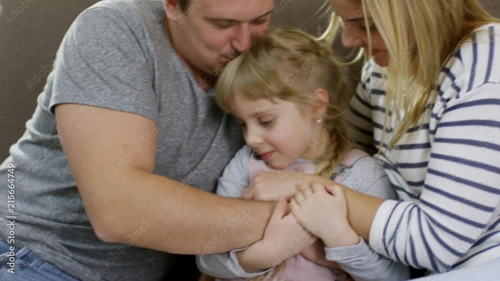 Loving mother and father embracing and caressing cute little daughter while bonding together on sofa at home Loving mother and father embracing and caressing cute little daughter while bonding together on sofa at home