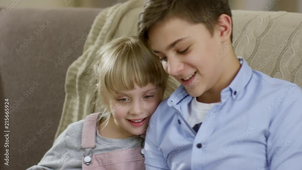 Cute little girl sitting together with elder brother of sofa, smiling and talking while reading a book Cute little girl sitting together with elder brother of sofa, smiling and talking while reading a book