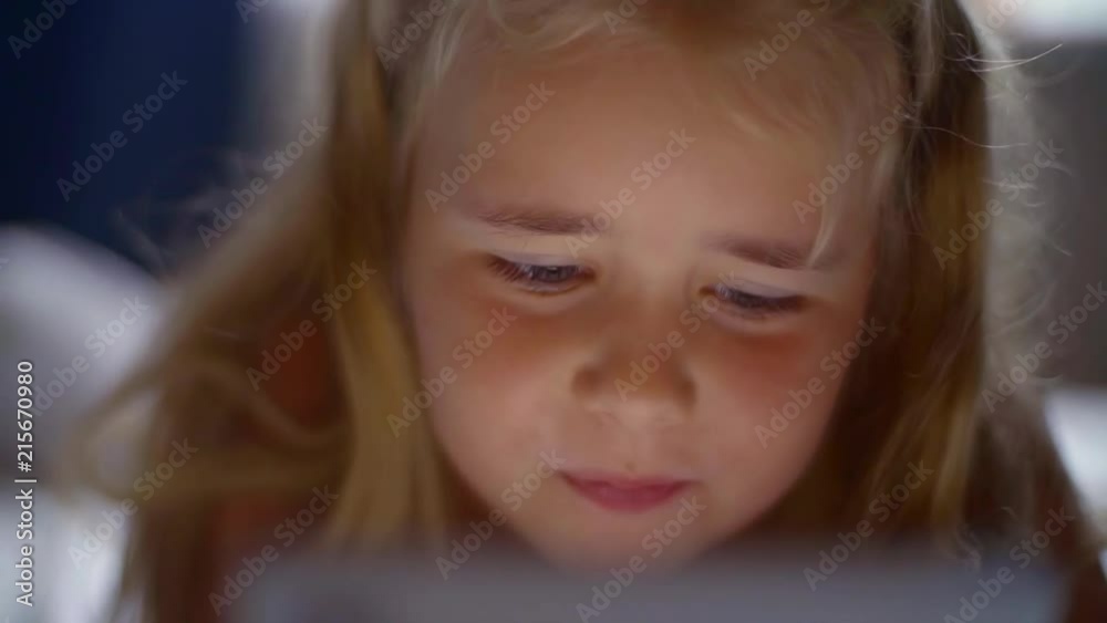 Happy smiling child lying in bed and enthusiastically playing the game on the smartphone. The child's face is illuminated by a bright monitor Happy smiling child lying in bed and enthusiastically playing the game on the smartphone. The child's face is illuminated by a bright monitor