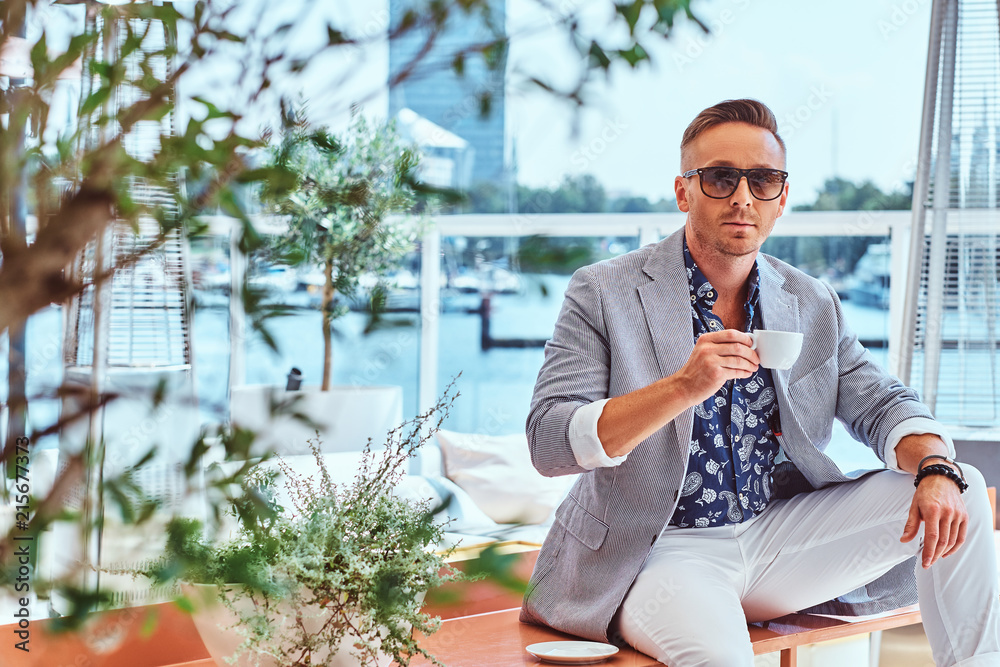 Confident successful man with stylish hair dressed in modern elegant clothes holds cup of coffee while sitting on a table at outdoor cafe against the background of city wharf. - obrazy, fototapety, plakaty 
