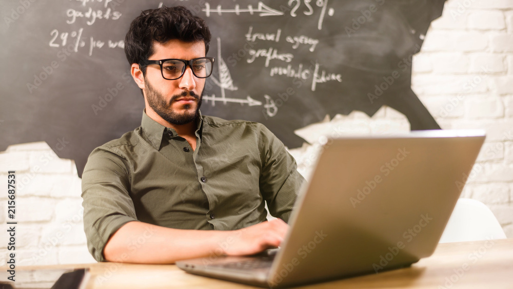 Attractive multicultural bearded businessman in glasses and green shirt working behind the laptop and writting some words in the office on the some diagrams background Attractive multicultural bearded businessman in glasses and green shirt working behind the laptop and writting some words in the office on the some diagrams background