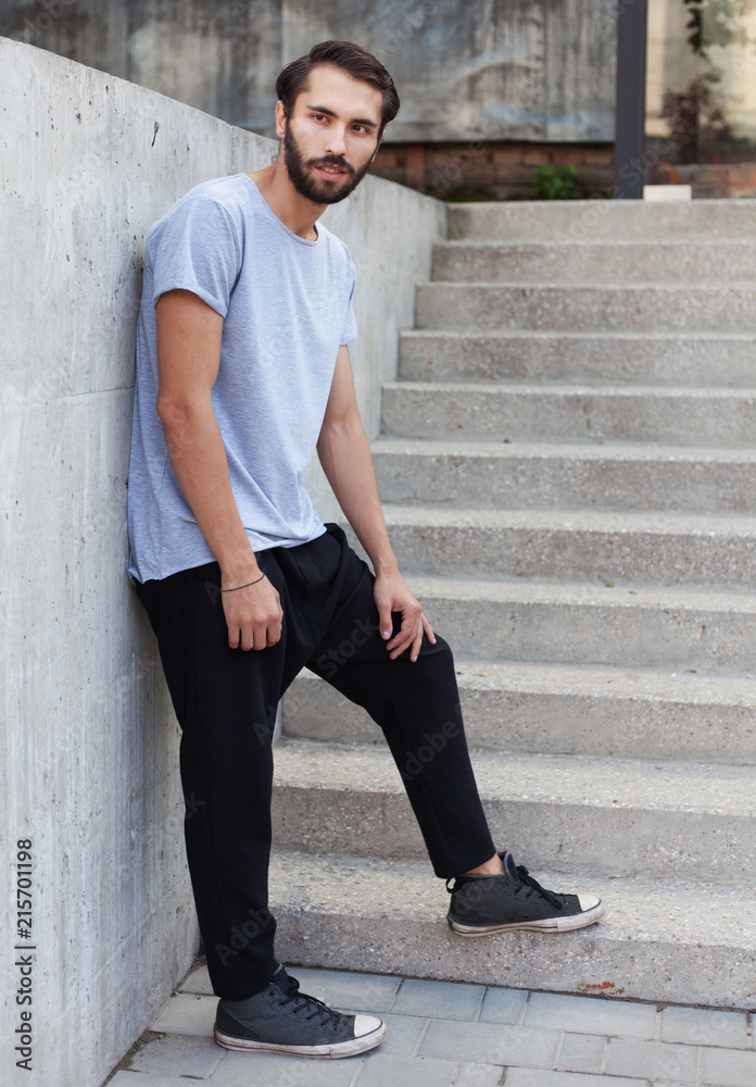 A bearded hipster man in a gray T-shirt, pants pants posing against a gray stone wall in the afternoon.