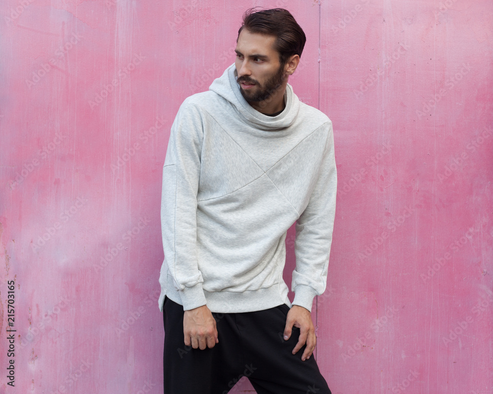 Men's youth street fashion. Gray hoodie and black pants on a young man posing against a pink wall background.
