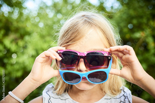 Child cute girl having several sunglasses on her face and doing fun during a hot summer day. Greenery background