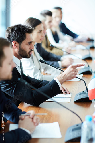 Aggressive confident handsome young bearded political leader in suit expressing his position and pointing with forefinger while speaking into microphone at international meeting photo