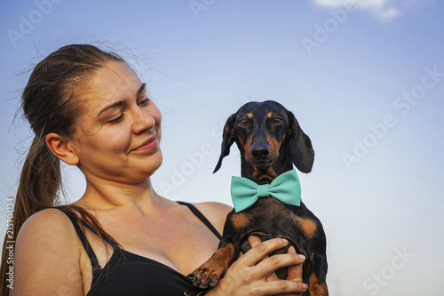 Portrait of a young beautiful girl and her dog dachshund in a blue bow, playing outdoor on the blue sky background