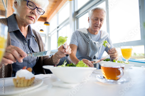 Low angle portrait of modern senior couple eating salad sitting at table in cafe