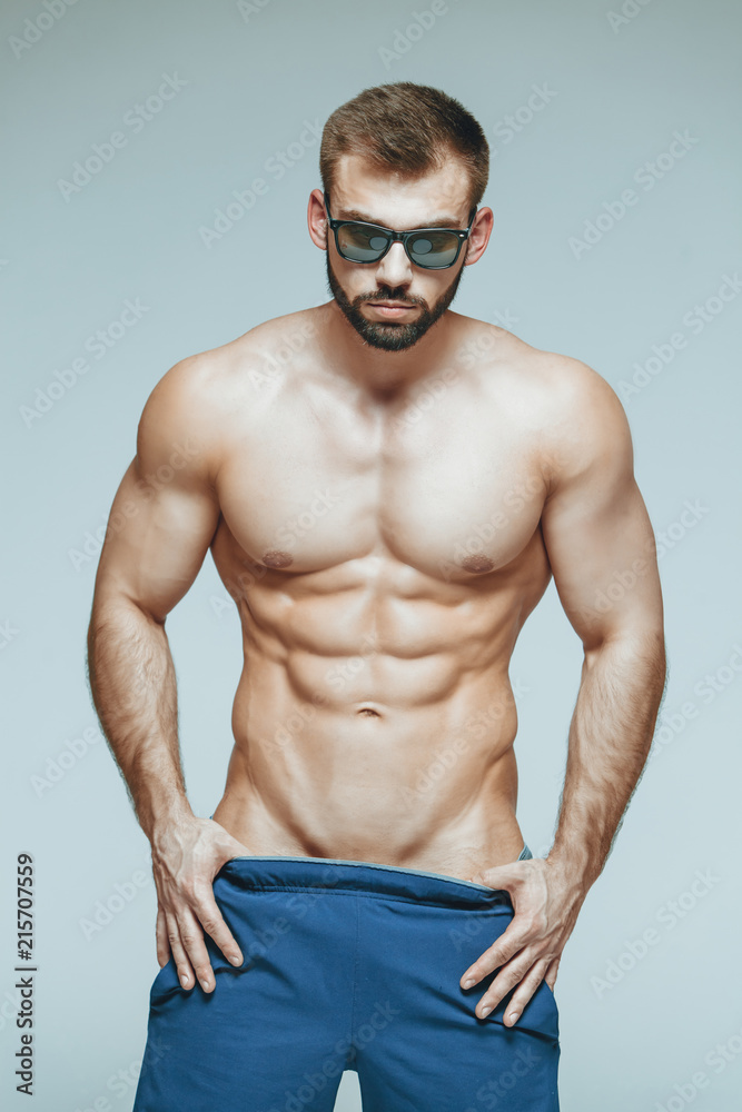 .bodybuilder posing. Beautiful sporty guy male power. Fitness muscled in blue shorts and sunglasses. on isolated grey background. Man with muscular torso. Strong Athletic Man Fitness Model Torso