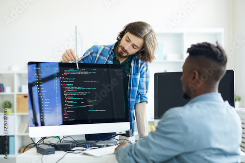Confident professional multiethnic coders discussing programming language: hipster young man pointing at screen and explaining programming algorithm