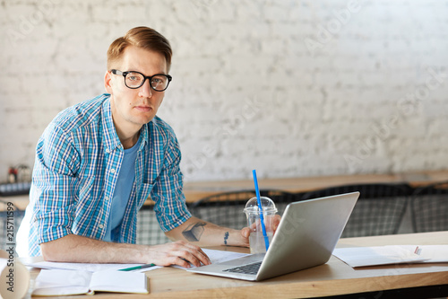 Portrait of modern young man wearing glasses and casual shirt looking at camera while working with laptop in office, copy space