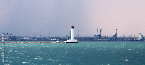 Lighthouse in the sea against the backdrop of a cargo port. Summer seascape with a white lighthouse with red top. Black Sea. Sea Port of Odessa. photo
