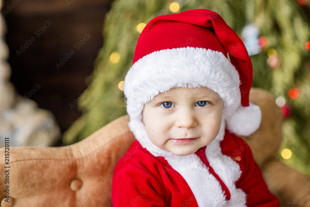 Close-up portrait of a Little boy dressed as a red Santa Claus, playing by a stone fireplace with bright garlands, putting a gift box under the Christmas tree. New Year card. The baby laughs, has fun. Close-up portrait of a Little boy dressed as a red Santa Claus, playing by a stone fireplace with bright garlands, putting a gift box under the Christmas tree. New Year card. The baby laughs, has fun.