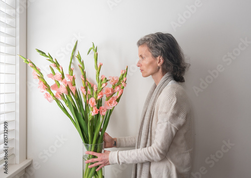 Vászonkép Middle aged woman with grey hair arranging pink gladioli in vase next to window