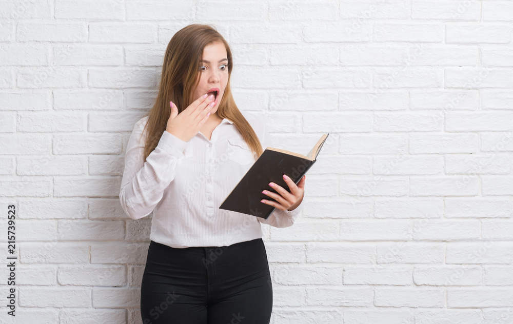 Young adult woman standing over white brick wall reading a book cover mouth with hand shocked with shame for mistake, expression of fear, scared in silence, secret concept Young adult woman standing over white brick wall reading a book cover mouth with hand shocked with shame for mistake, expression of fear, scared in silence, secret concept