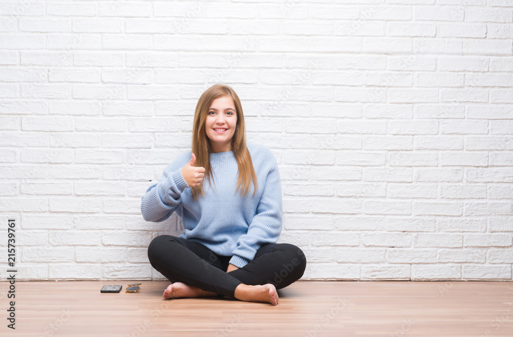 Young adult woman sitting on the floor in autumn over white brick wall doing happy thumbs up gesture with hand. Approving expression looking at the camera with showing success. Young adult woman sitting on the floor in autumn over white brick wall doing happy thumbs up gesture with hand. Approving expression looking at the camera with showing success.