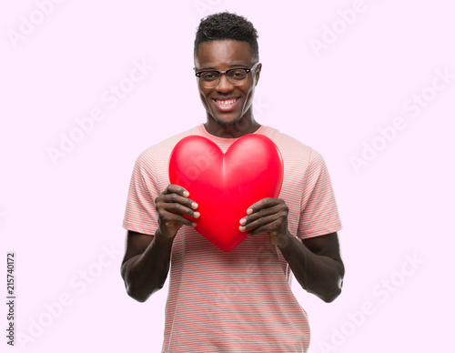 Young african american man holding red heart with a happy face standing and smiling with a confident smile showing teeth © Krakenimages.com