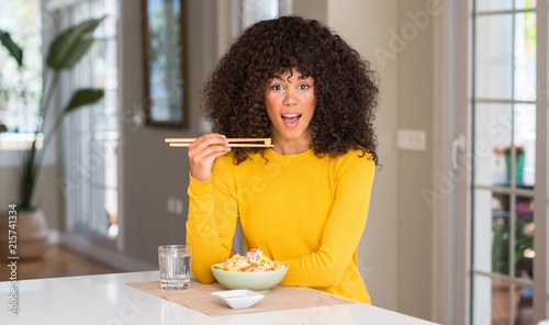 African american woman eating asian rice at home scared in shock with a surprise face, afraid and excited with fear expression